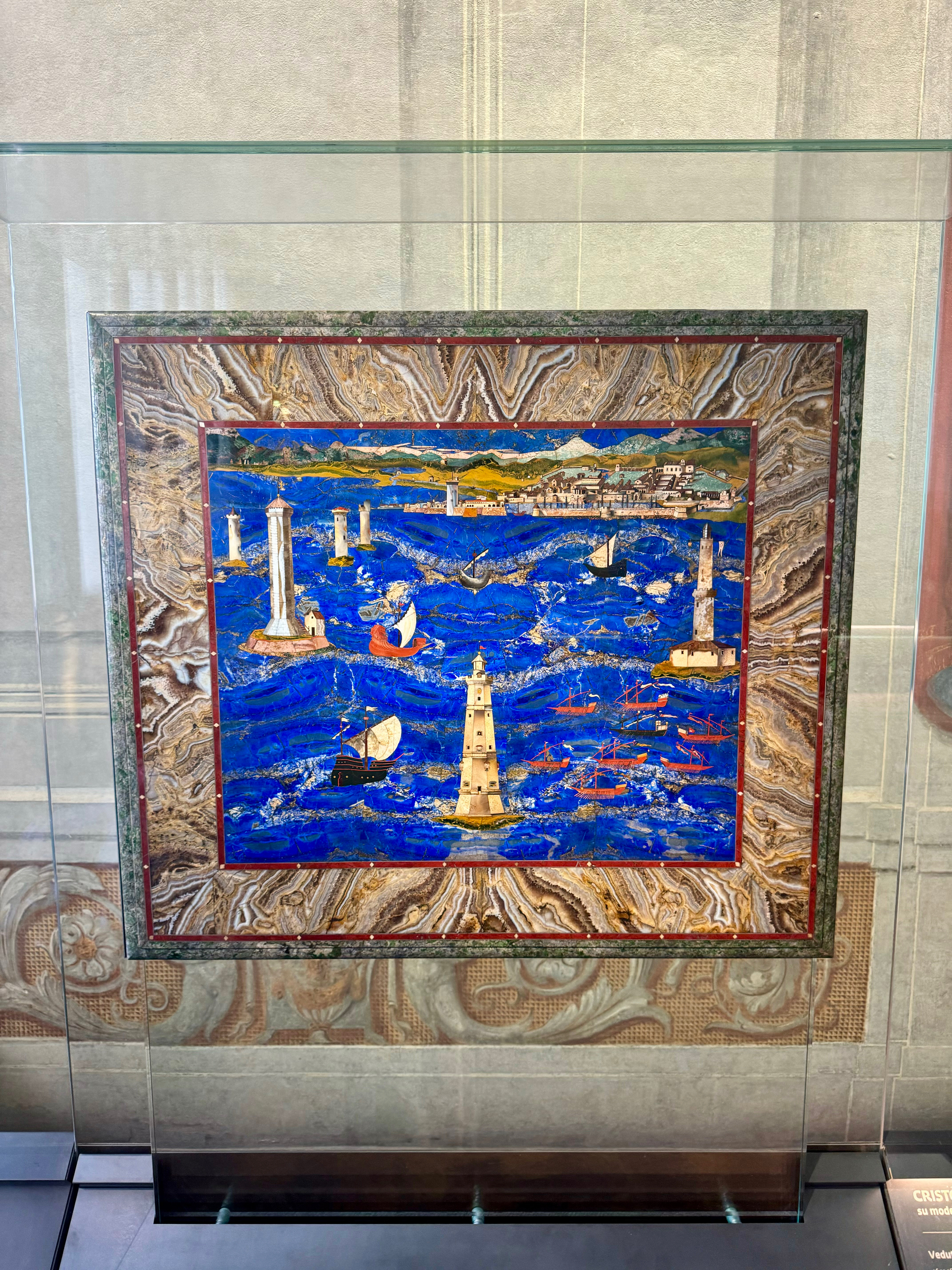 This image shows a colorful artwork display featuring a vivid depiction of a coastal scene with a tall lighthouse and several sailboats on a vibrant blue sea. The artwork is encased in a glass display with a decorative, patterned stone frame.