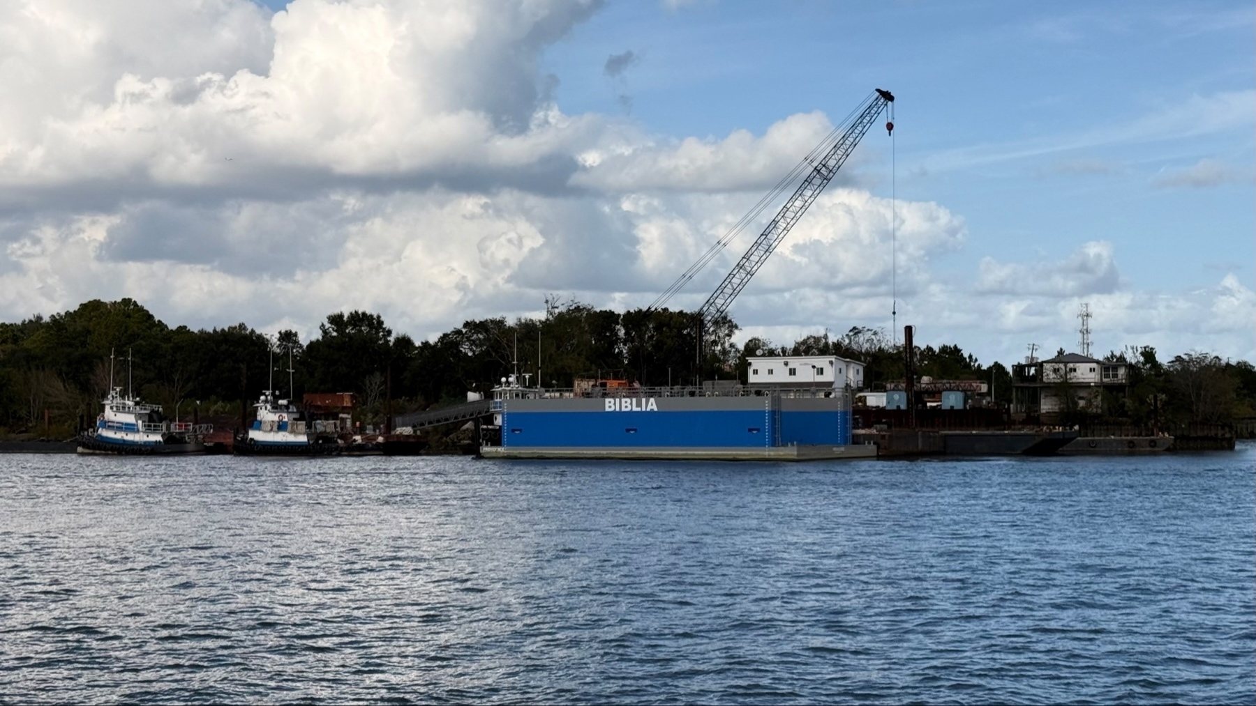 A tranquil waterfront scene featuring two small tugboats and a large blue barge labeled “BIBLIA,” equipped with a crane. The background contains trees and industrial buildings under a partly cloudy sky.