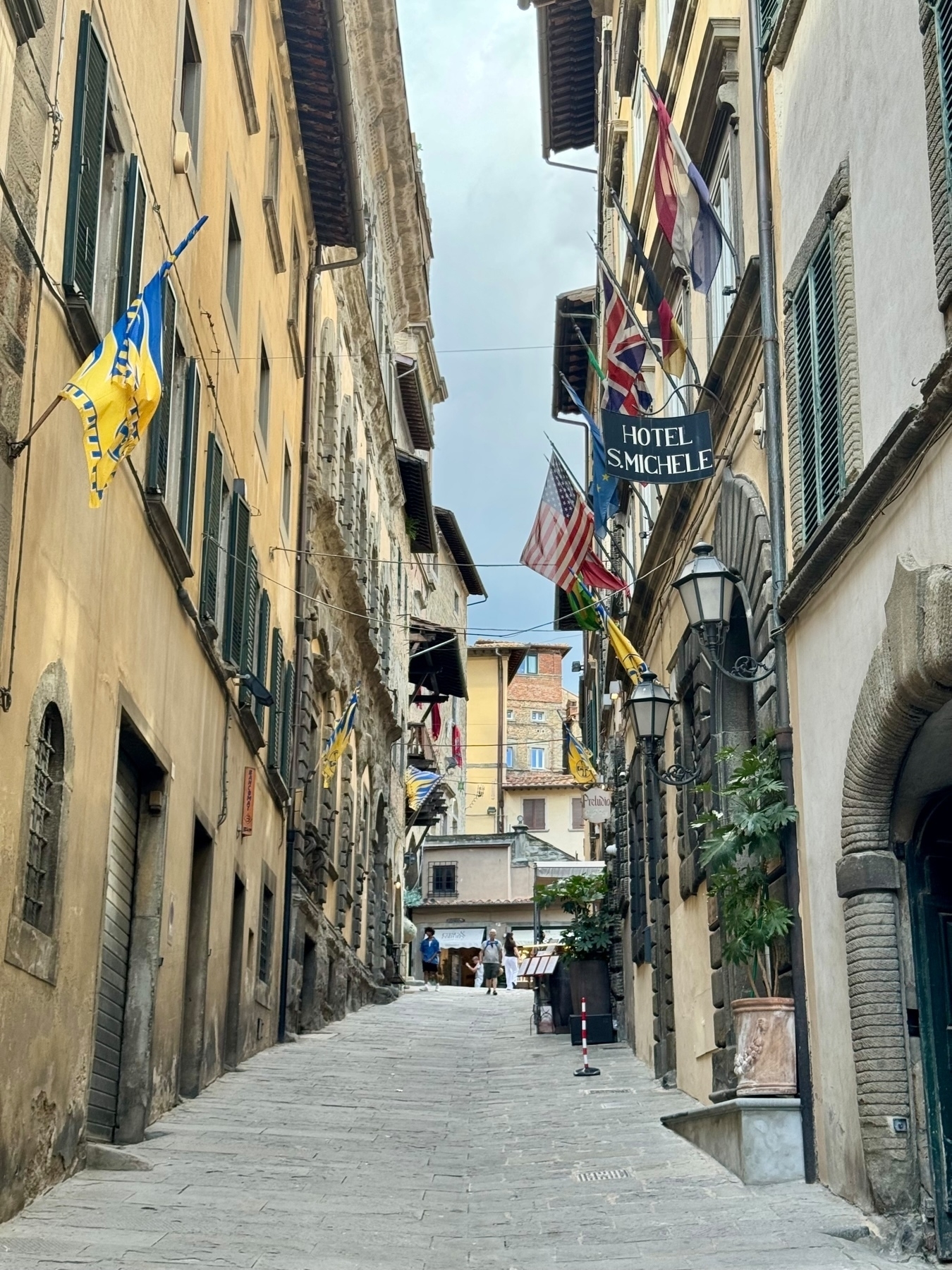 A narrow, cobbled street lined with historic buildings and colorful flags hanging from windows, with a sign for “Hotel S. Michele” on the right side. A few pedestrians can be seen in the distance. 