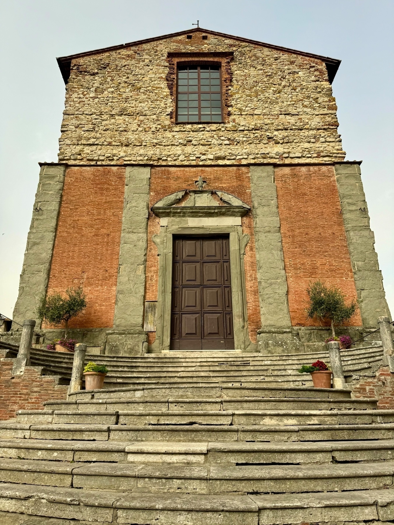 A stone and brick church facade with a large wooden door and a tall window above it. The church is accessed via a set of wide, stone stairs. Two potted plants flank the steps, and two additional potted plants are placed higher up. 