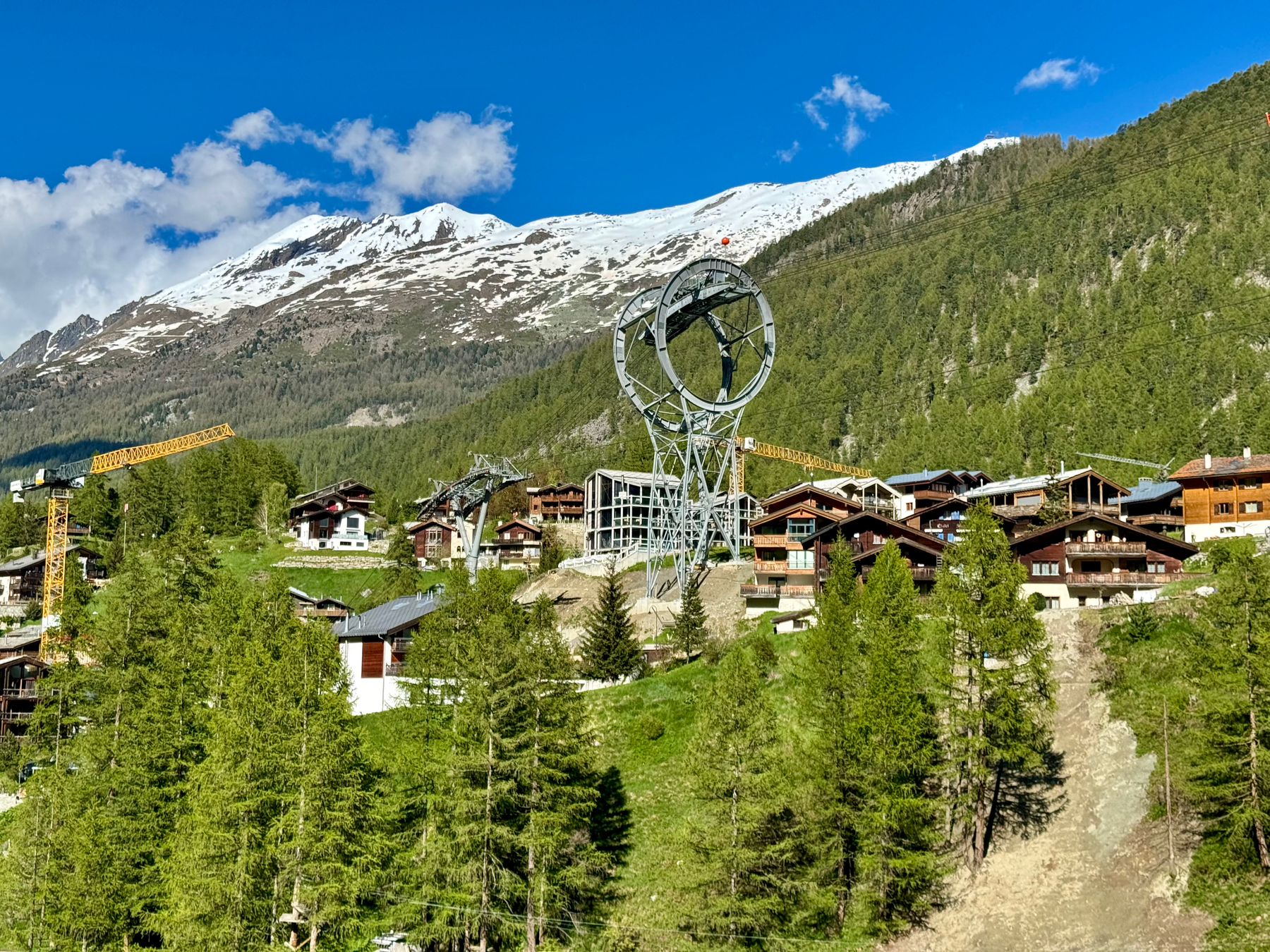 Alpine village with chalet-style buildings, surrounded by forest, with a large metal structure in the foreground and snow-capped mountains in the background.
