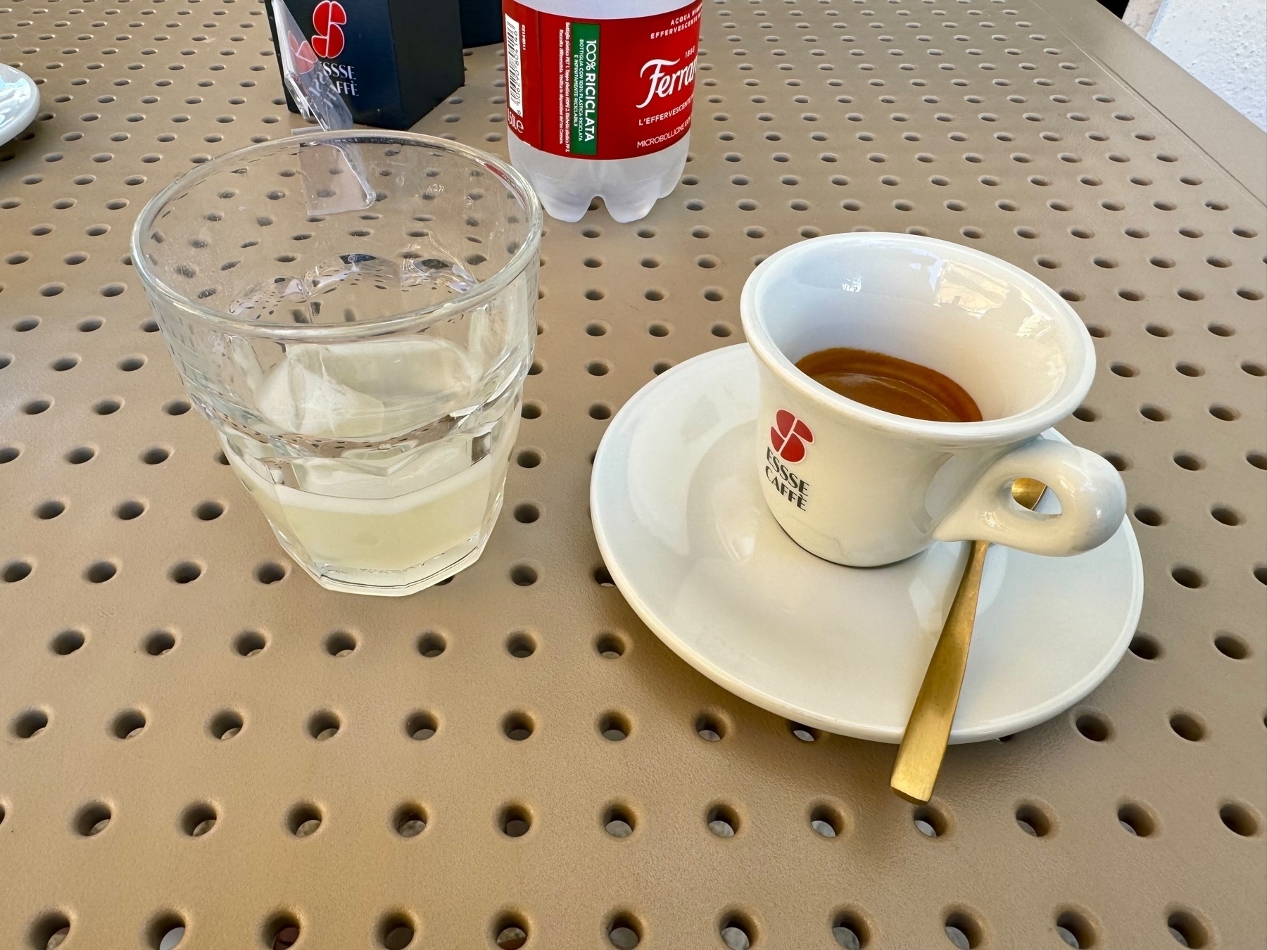 The image shows a glass of almond syrup with ice, a cup of espresso on a saucer with a spoon, and a bottle of water labeled “Ferrarelle.” They are placed on a beige perforated table.