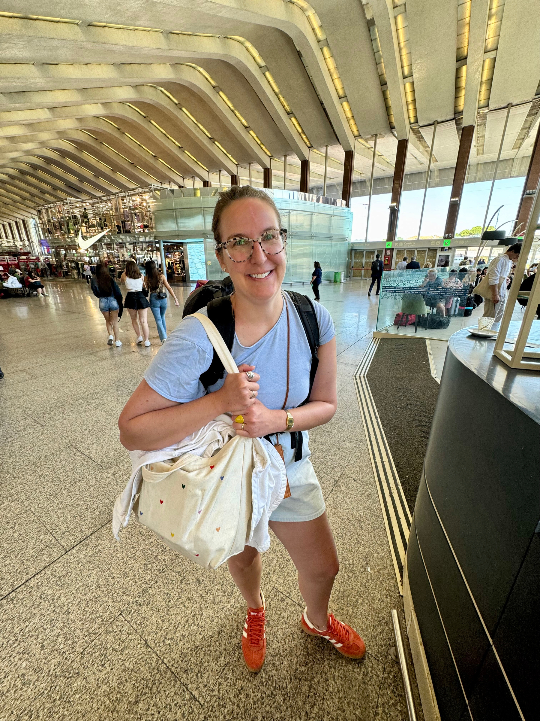 A woman is standing in a train terminal, smiling at the camera. She is holding a tote bag with small heart designs and wearing a light blue T-shirt, beige shorts, red sneakers, glasses, and a wristwatch.