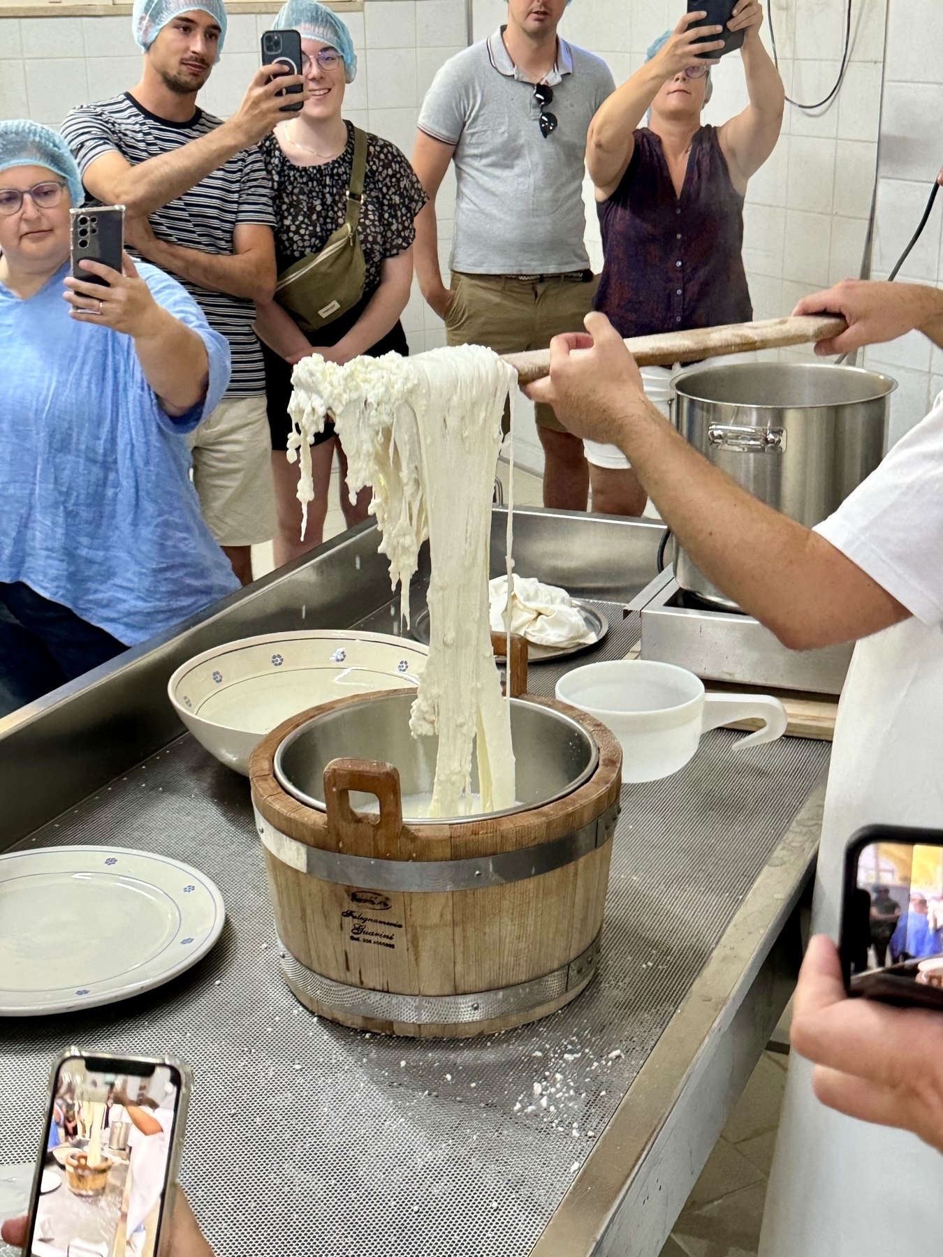 A group of people wearing hairnets are observing and capturing photos and videos of a cheese-making process. A person is lifting a large, stretchy mass of curd from a wooden vat using a long wooden stick.