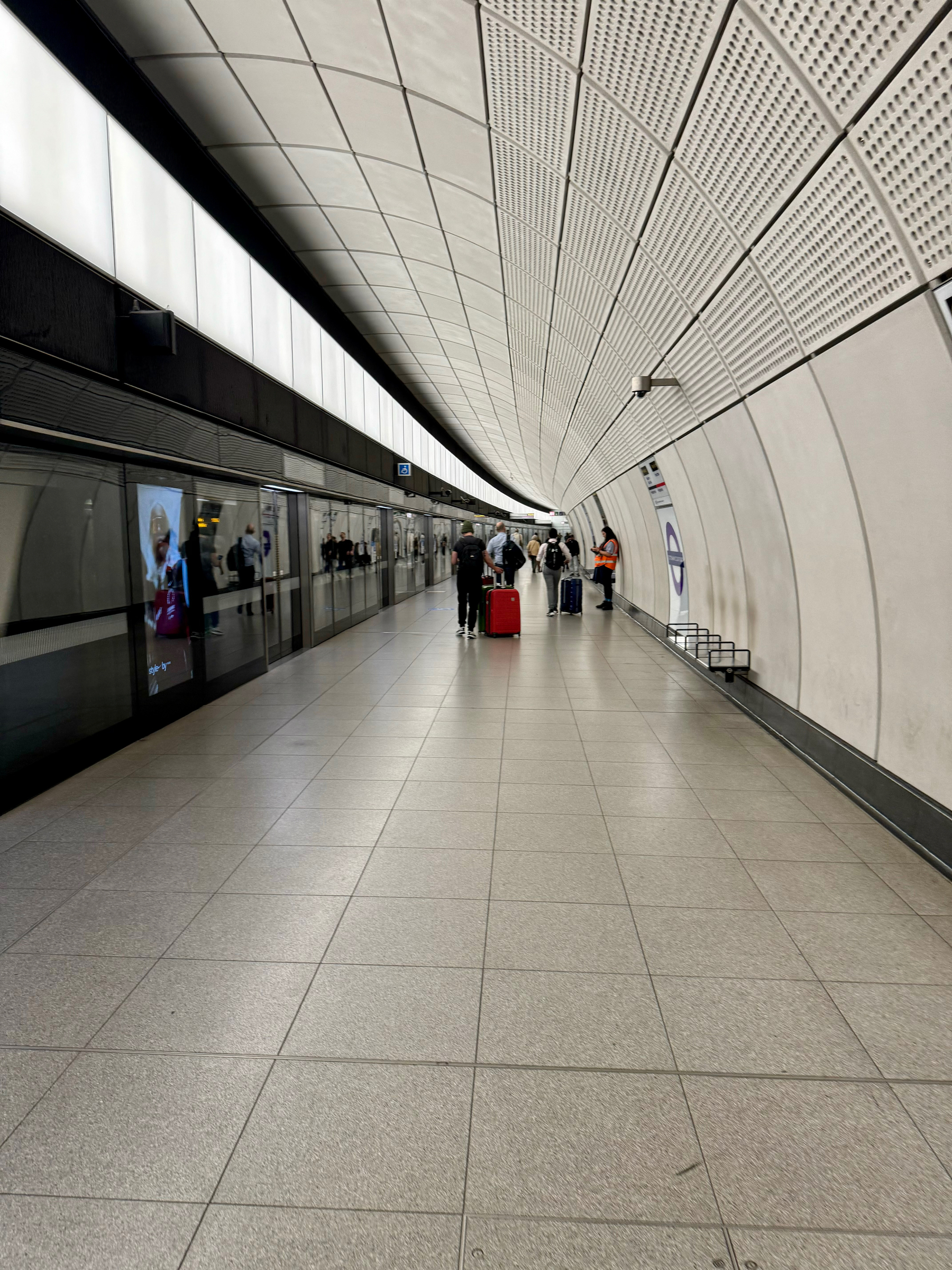 A subway station on the Elizabeth line from the airport. Interior of a modern subway station with curved walls and ceiling, featuring a reflective surface on one side and a tiled flooring leading to platform gates where people are standing and walking. One individual is noticeable in the foreground pulling a red suitcase.