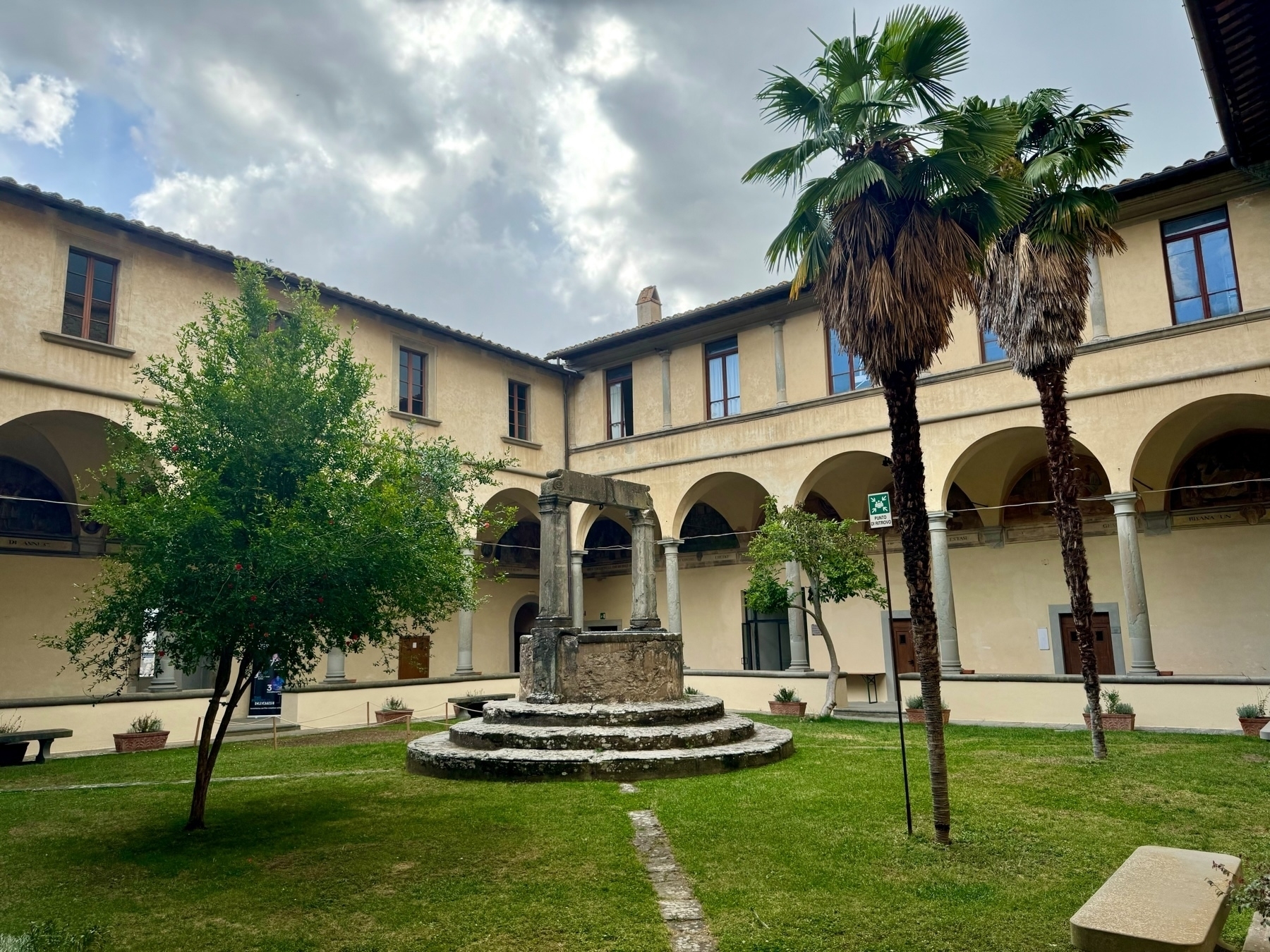 A peaceful courtyard featuring a central stone well surrounded by a grassy area with a tree and two tall palms. The courtyard is bordered by a two-story building with arched walkways and multiple windows. The sky above is partly cloudy.