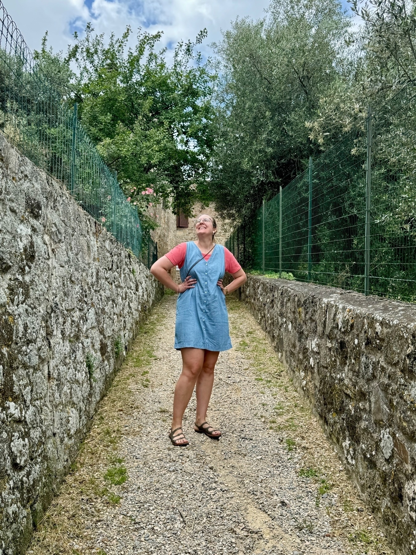 A person stands in a narrow alleyway lined with stone walls and metal fences, looking upwards and smiling. They are wearing a denim dress over a red shirt and sandals. The alley is surrounded by greenery and trees, and a partly cloudy sky is visible. 