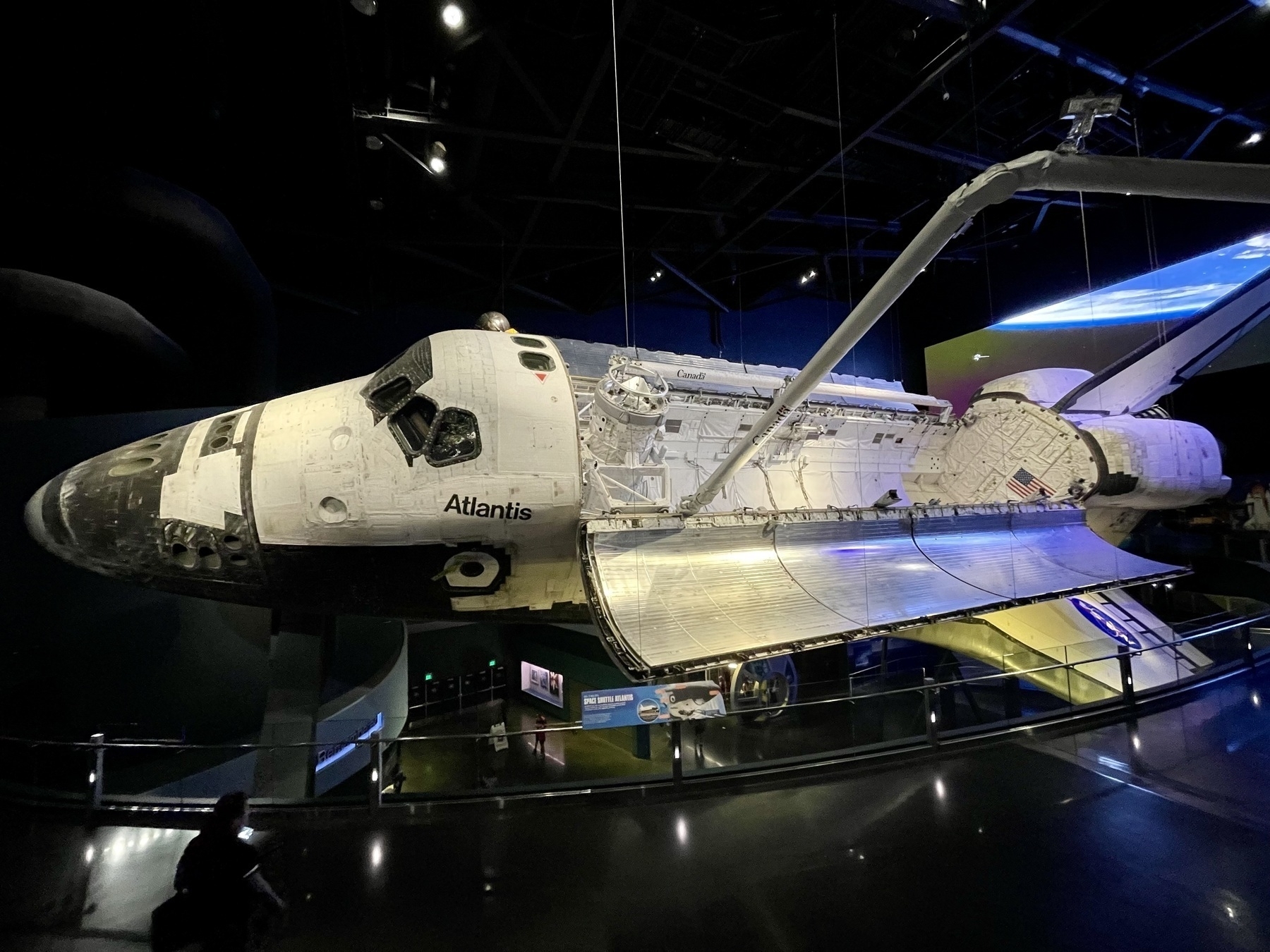 The image shows the Space Shuttle Atlantis on display in a museum setting. The shuttle is suspended with its cargo bay doors open and robotic arm extended. Dim lighting and informational displays are visible in the background.