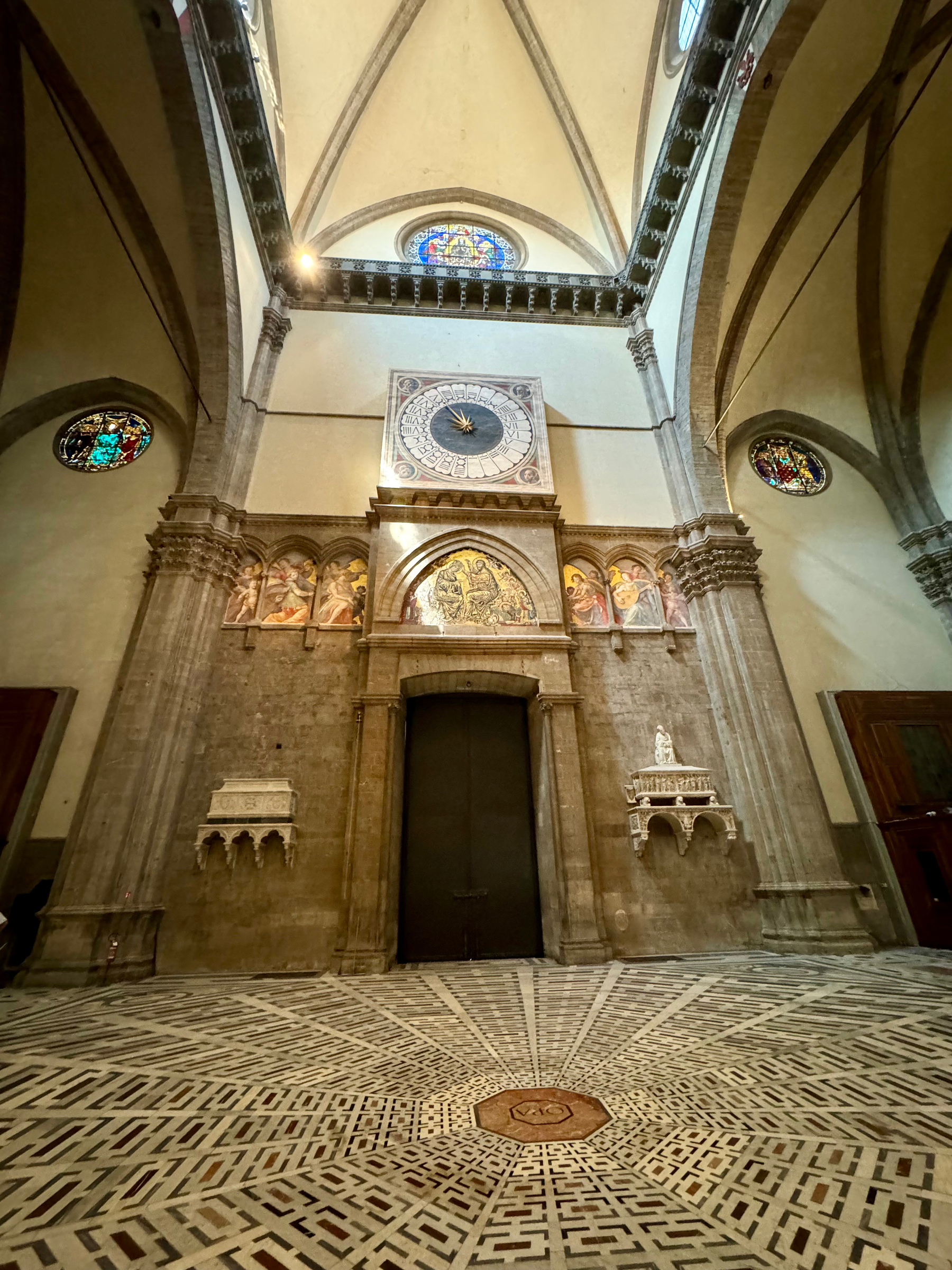 The image shows the interior of a historic cathedral, featuring a large ornate clock on the wall above a dark wooden door, flanked by intricate stone carvings and religious frescoes. The ceiling is vaulted with stained glass windows above. The patterned marble floor. 