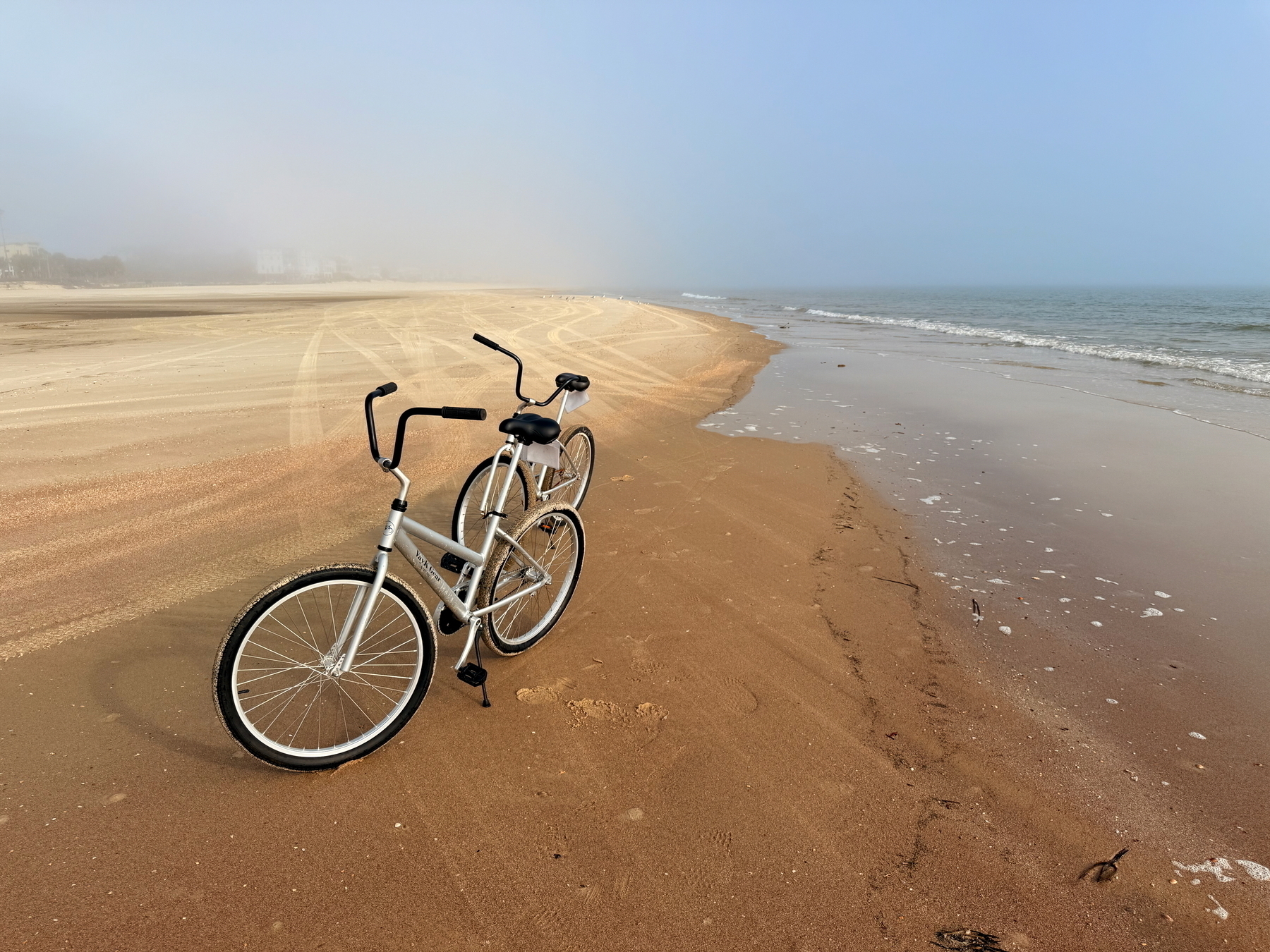 Auto-generated description: Two bicycles are parked on a sandy beach near the ocean on a foggy day.