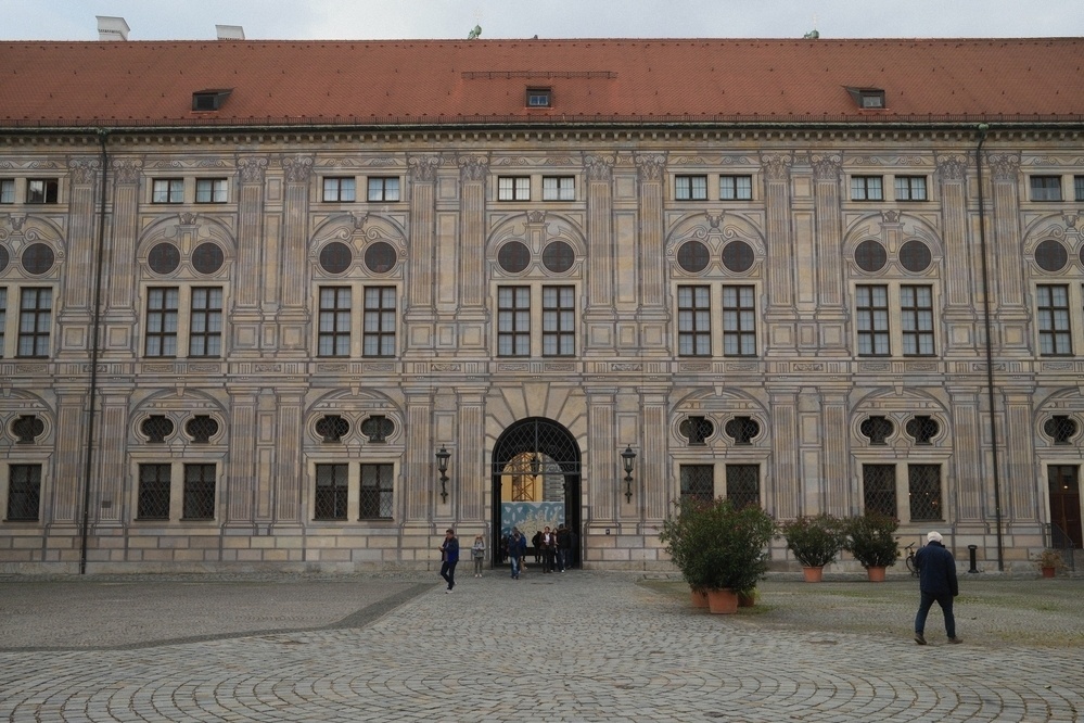 A large, ornate building features symmetrical windows and decorative patterns on its facade, with people walking in front of it.