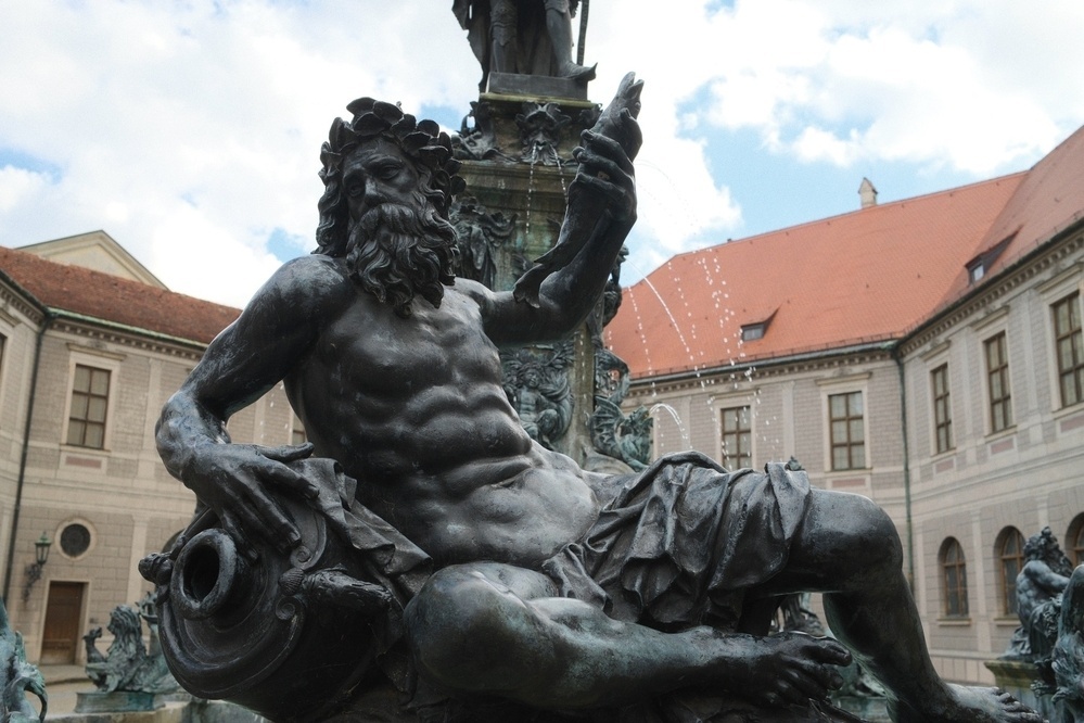 A statue of a reclining bearded figure with muscular features holding a cornucopia is part of an ornate fountain set against the backdrop of a historic building.