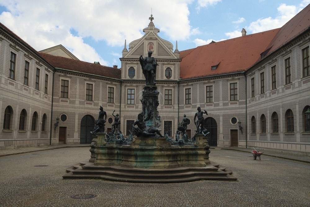 A historic courtyard features an ornate fountain with multiple statues, surrounded by an elegant building with arched windows and a red-tiled roof.