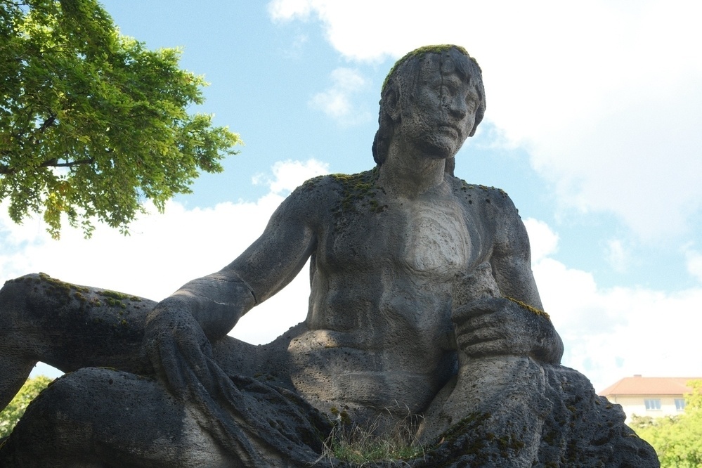A stone statue of a reclining figure with an expression of calm is set against a backdrop of trees and sky.