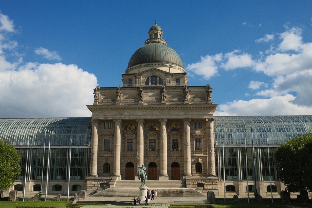 A grand historic building with a large dome and columned facade is surrounded by a glass structure, set against a blue sky with clouds.