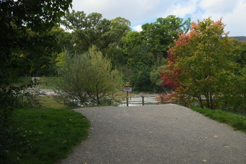 A pathway in a park leads to a lake, surrounded by lush green and autumn-colored trees under a partly cloudy sky.