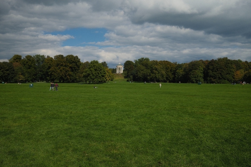 A wide expanse of green grass is surrounded by trees, with a small structure visible in the distance under a cloudy sky.