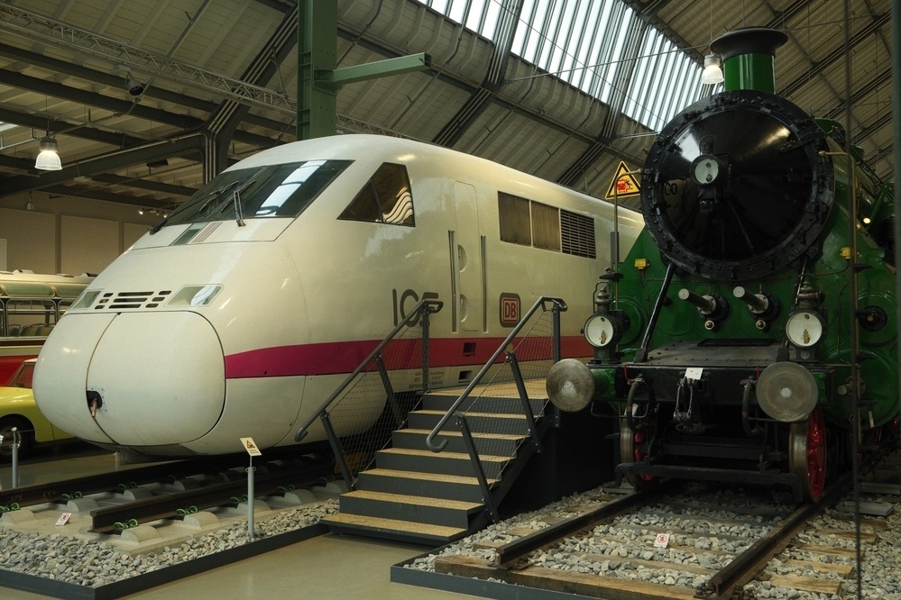 A modern high-speed train engine and a classic steam locomotive are displayed side by side in a museum setting.