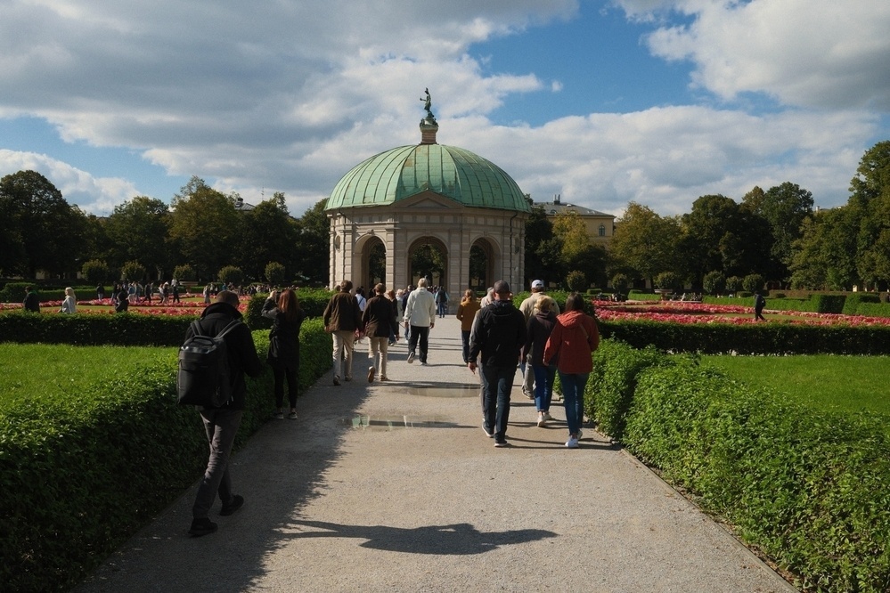 People are walking along a path toward a domed pavilion surrounded by gardens in a park.
