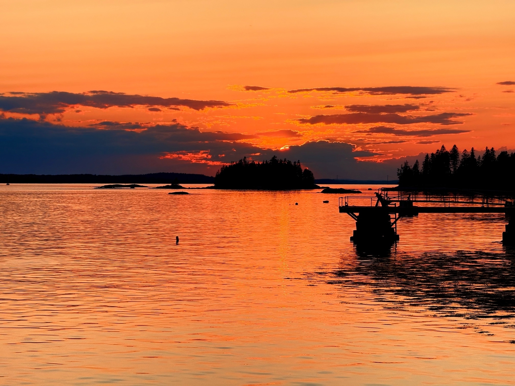 A vibrant sunset over a calm body of water with silhouettes of a dock and trees in the foreground. The sky is illuminated with shades of orange and red, reflecting beautifully on the water’s surface.