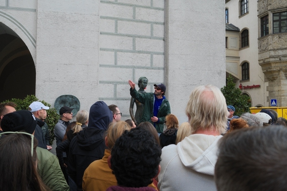 A group of people, led by a guide, gathers around a statue in an urban area with historical architecture.