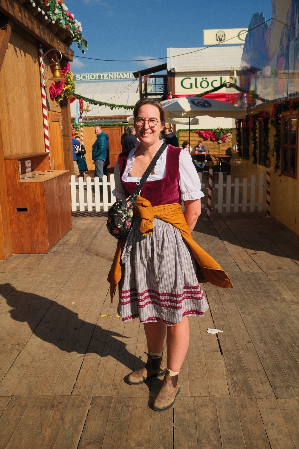 A person wearing traditional Bavarian clothing stands smiling in a festive outdoor setting with wooden structures and decorative elements.