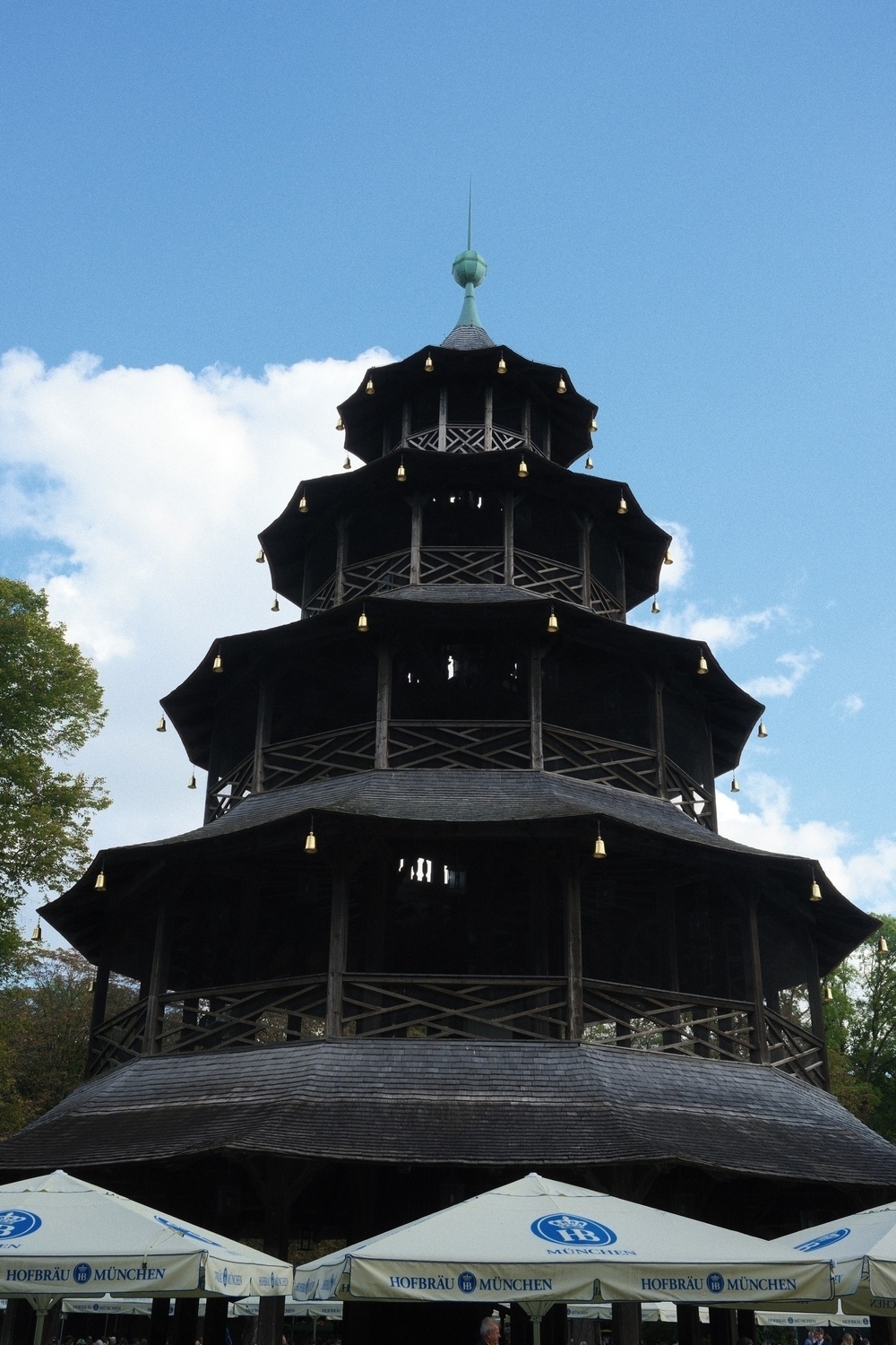 A multi-tiered, pagoda-style wooden structure stands against a blue sky with trees nearby and umbrellas in the foreground.