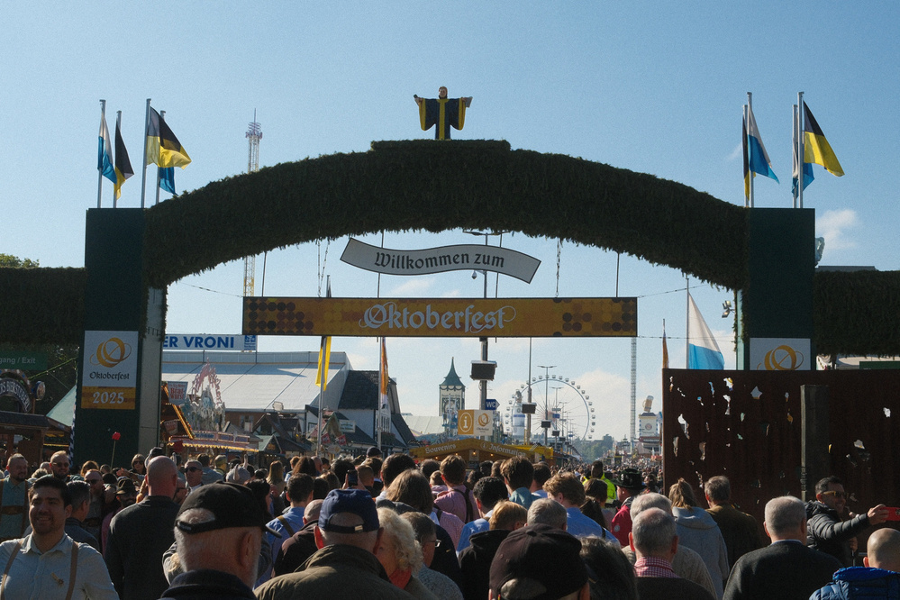 A large crowd gathers under an archway decorated with flags and the sign Oktoberfest at a lively festival.