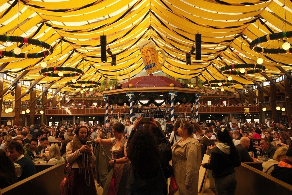 A large crowd enjoys a festive atmosphere beneath a yellow tent roof, with beer and decorations prominently featuring in the scene.