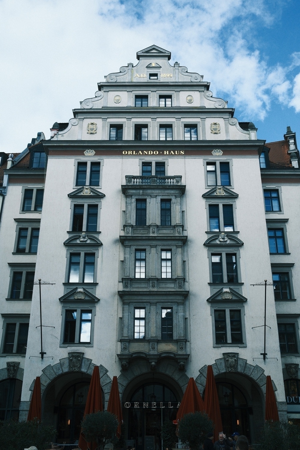 A historic multi-story building with ornate architecture features a central arched entrance, multiple windows, balconies, and a sign reading Orlando-Haus near the top, against a backdrop of blue sky.