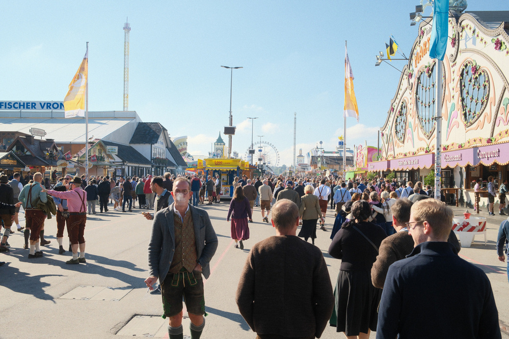 A large crowd of people, many wearing traditional Bavarian attire, are walking through a festive outdoor event with amusement rides and colorful structures.
