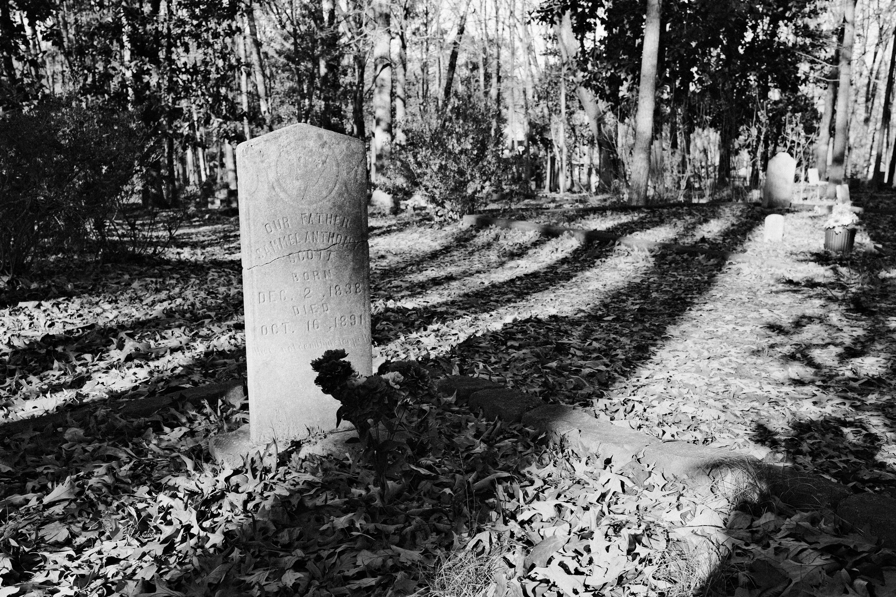 A black and white image of a graveyard with a tombstone in the foreground. The tombstone is inscribed with the name “Samuel Anthony Scott,” showing birth and death dates as December 2, 1838, and October 16, 1891.