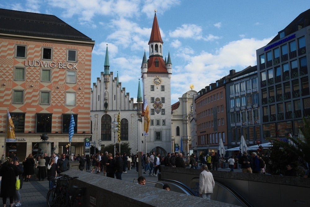 A bustling square features historic buildings, including a clock tower, surrounded by people and shops.