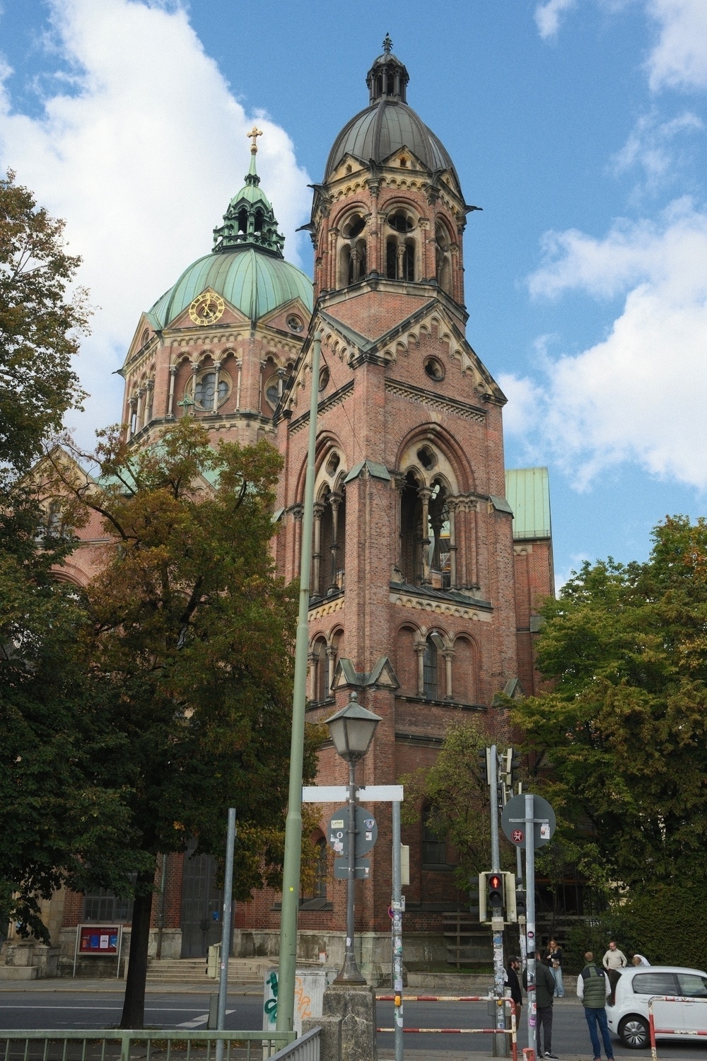 A large, historic brick church with prominent towers and green domes is surrounded by trees and a few people on the street.