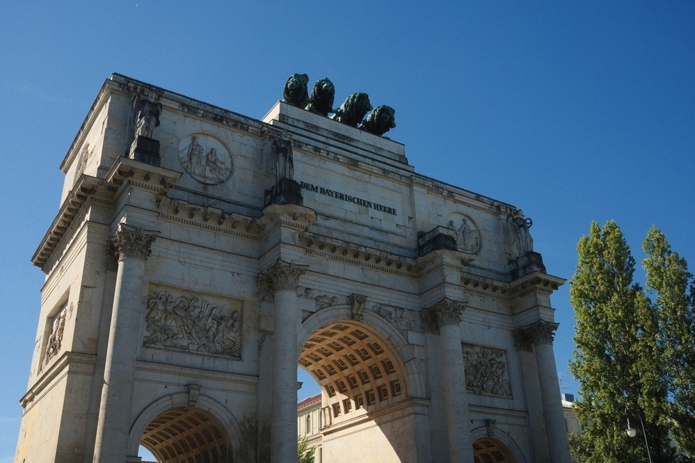A large triumphal arch with detailed carvings and sculptures sits against a clear blue sky, with trees visible to the right.