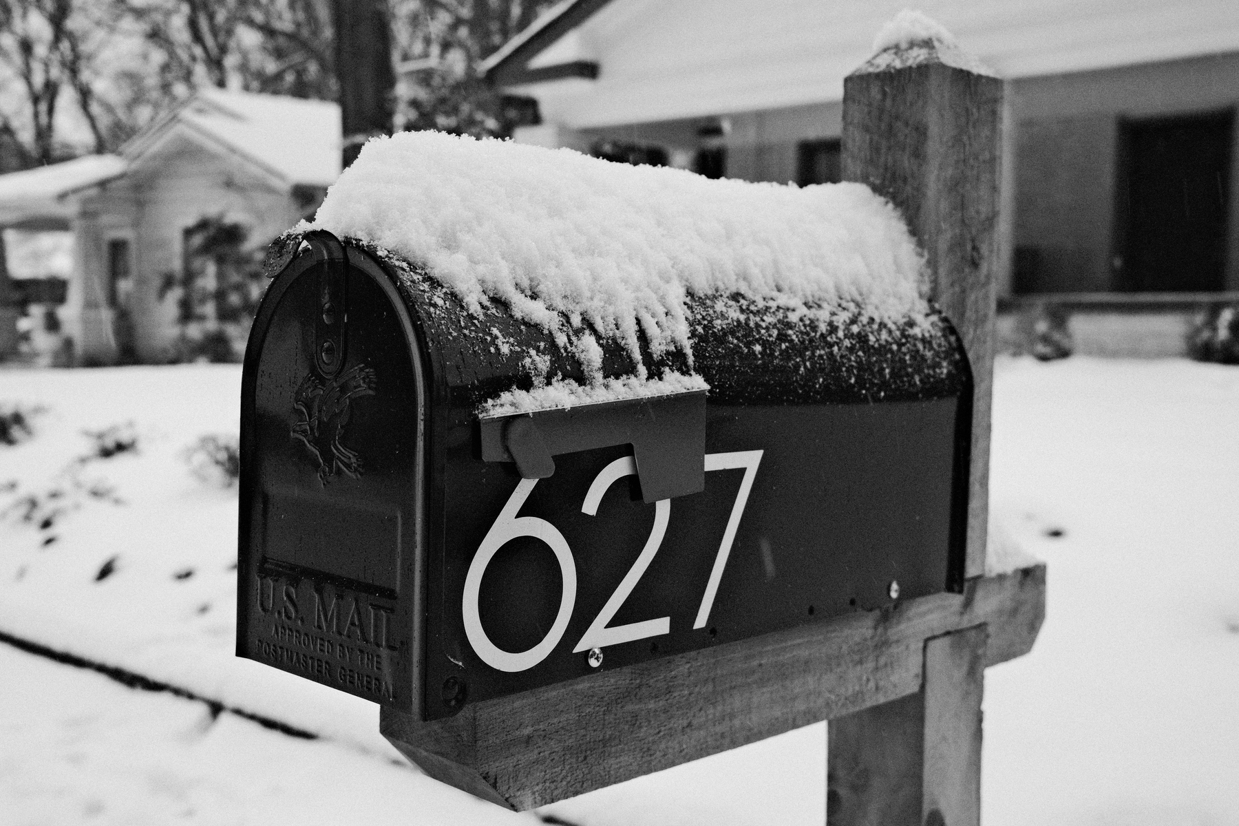 Auto-generated description: A snow-covered mailbox with the number 627 is mounted on a wooden post in front of snow-covered houses.