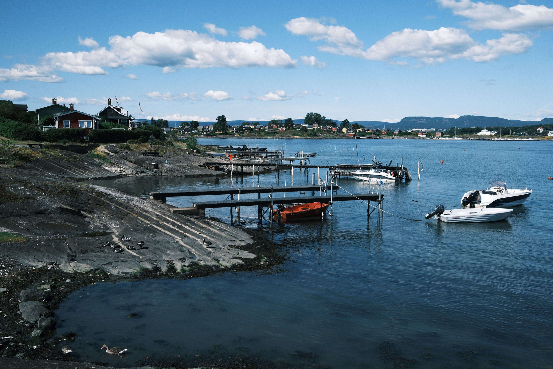 Auto-generated description: A peaceful waterfront scene features several small docks with boats moored, calm blue water, houses on the shore, and a partly cloudy sky.