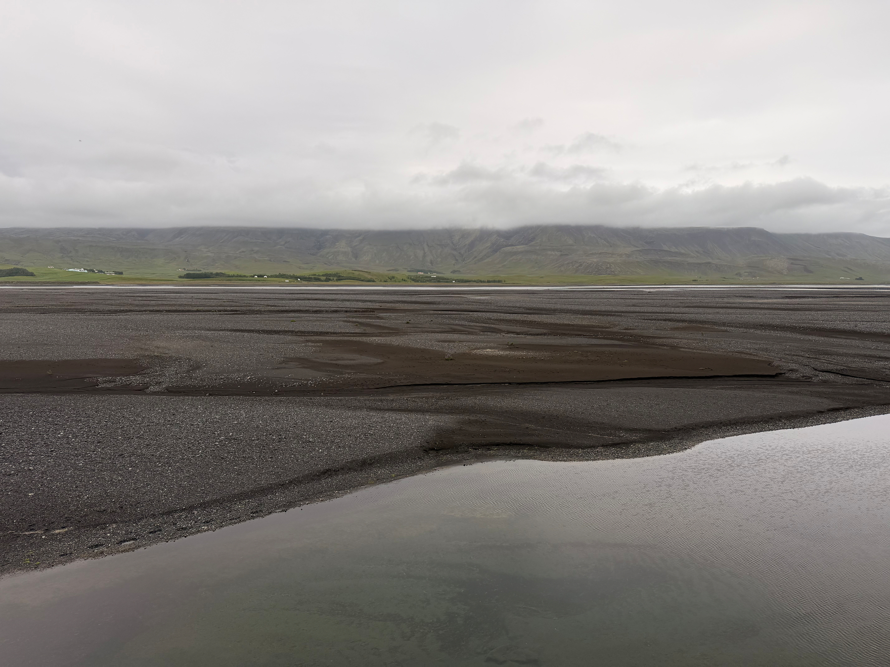 A calm body of water is bordered by dark sandy shores with green hills and a cloudy sky in the background.