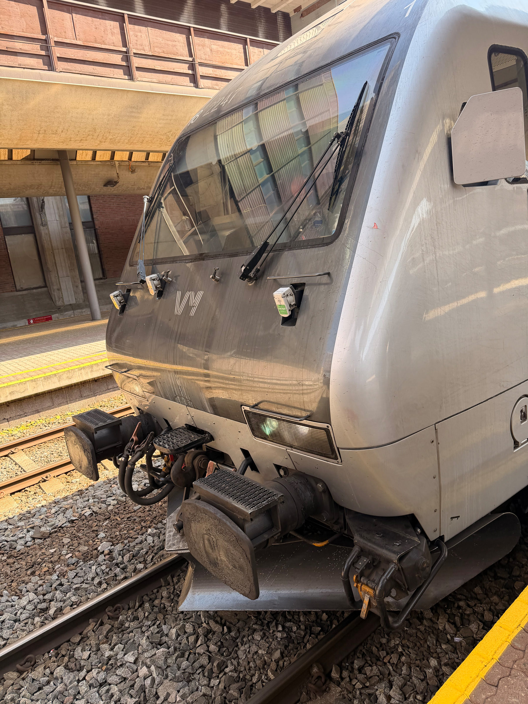 Auto-generated description: A modern silver train is stationed at a platform with visible tracks and part of a station structure in the background.