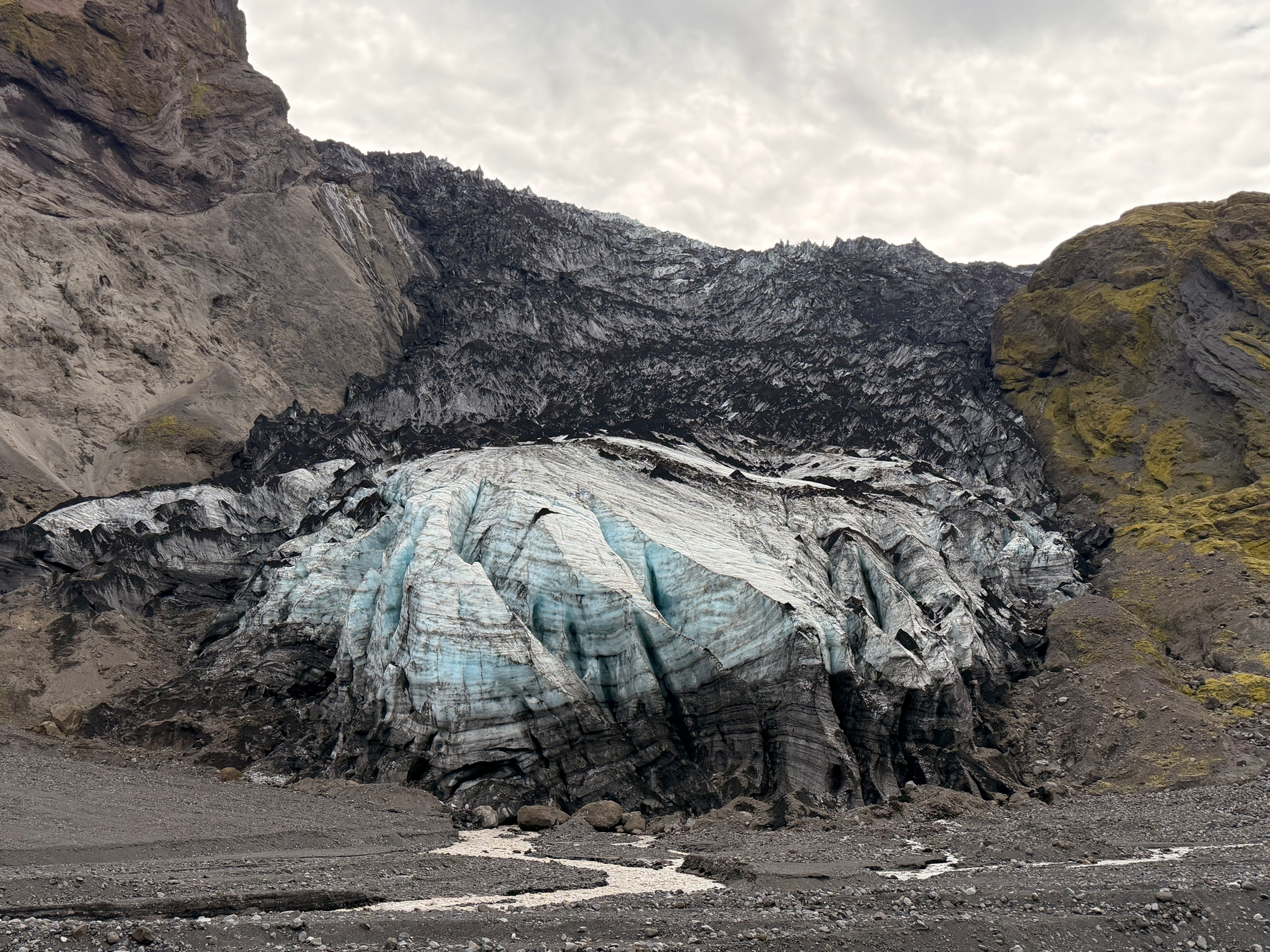 A partially melted glacier with visible blue ice and dark layers of volcanic ash is nestled between rocky cliffs under a cloudy sky.