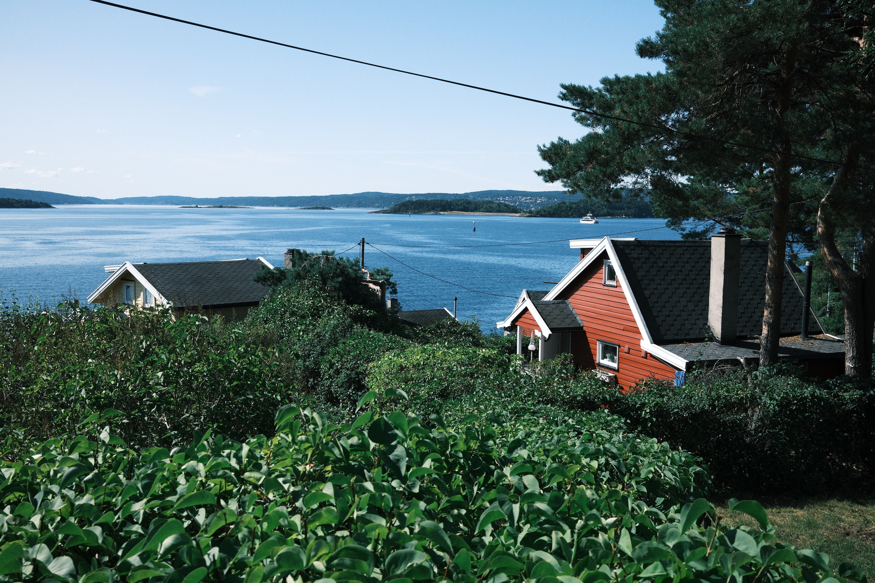 Auto-generated description: A scenic lakeside view features two houses surrounded by greenery, with a calm body of water and distant hills under a clear blue sky.