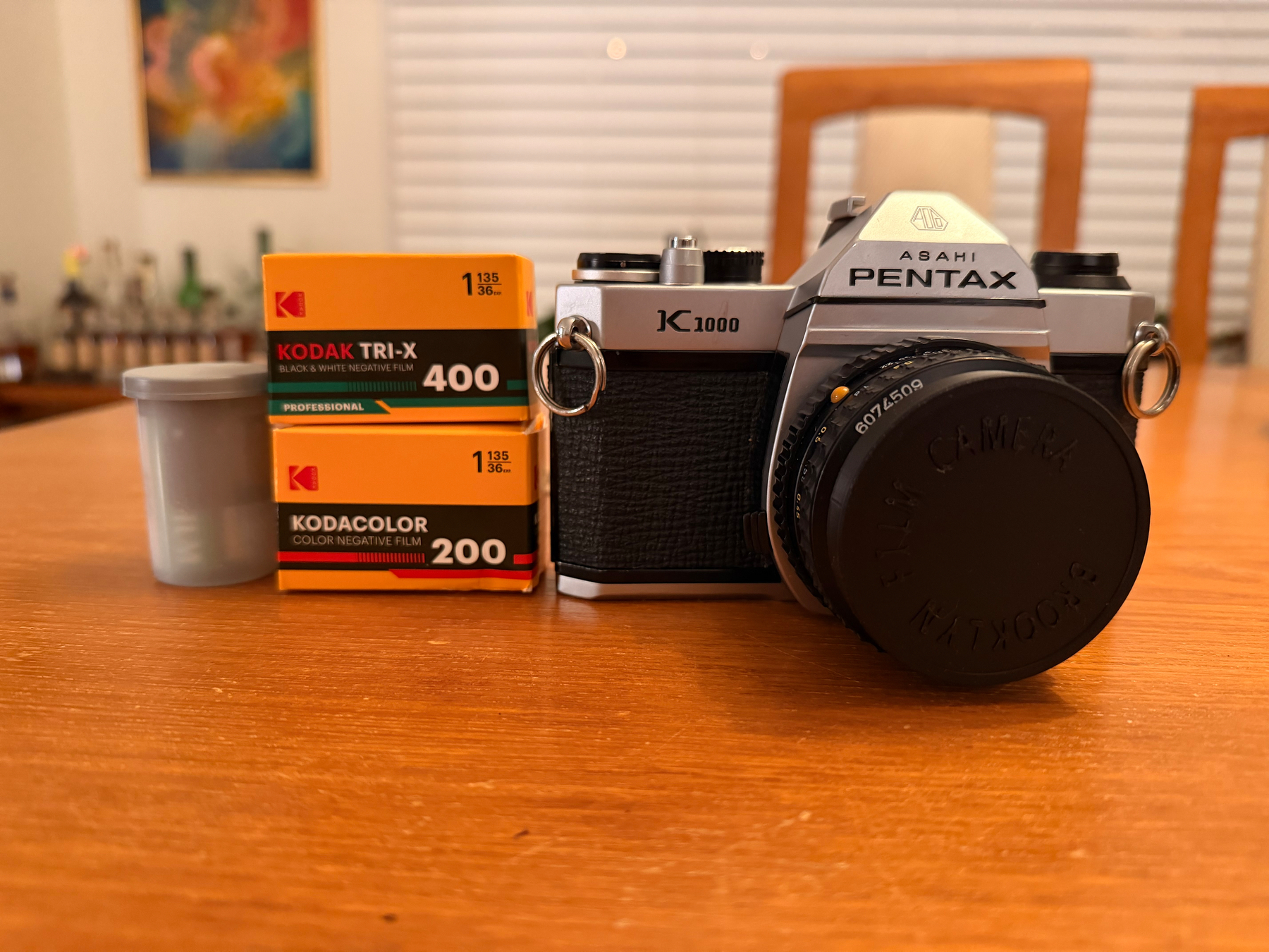 A Pentax K1000 camera is placed on a wooden table alongside two boxes of Kodak film and a film canister.