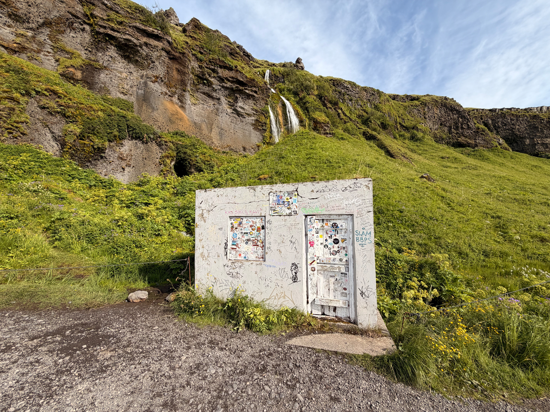 A small concrete structure covered in stickers and graffiti stands in front of a hillside with greenery and a waterfall cascading down the rocky cliff.
