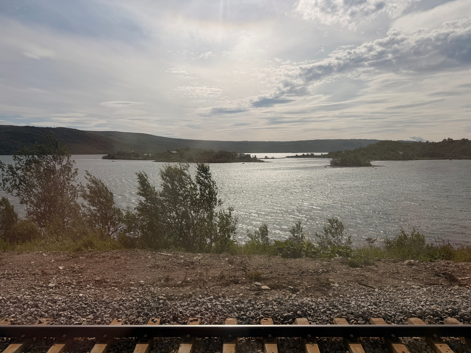 Auto-generated description: A peaceful lakeside scene is captured with calm water, distant hills, and railway tracks in the foreground under a partly cloudy sky.