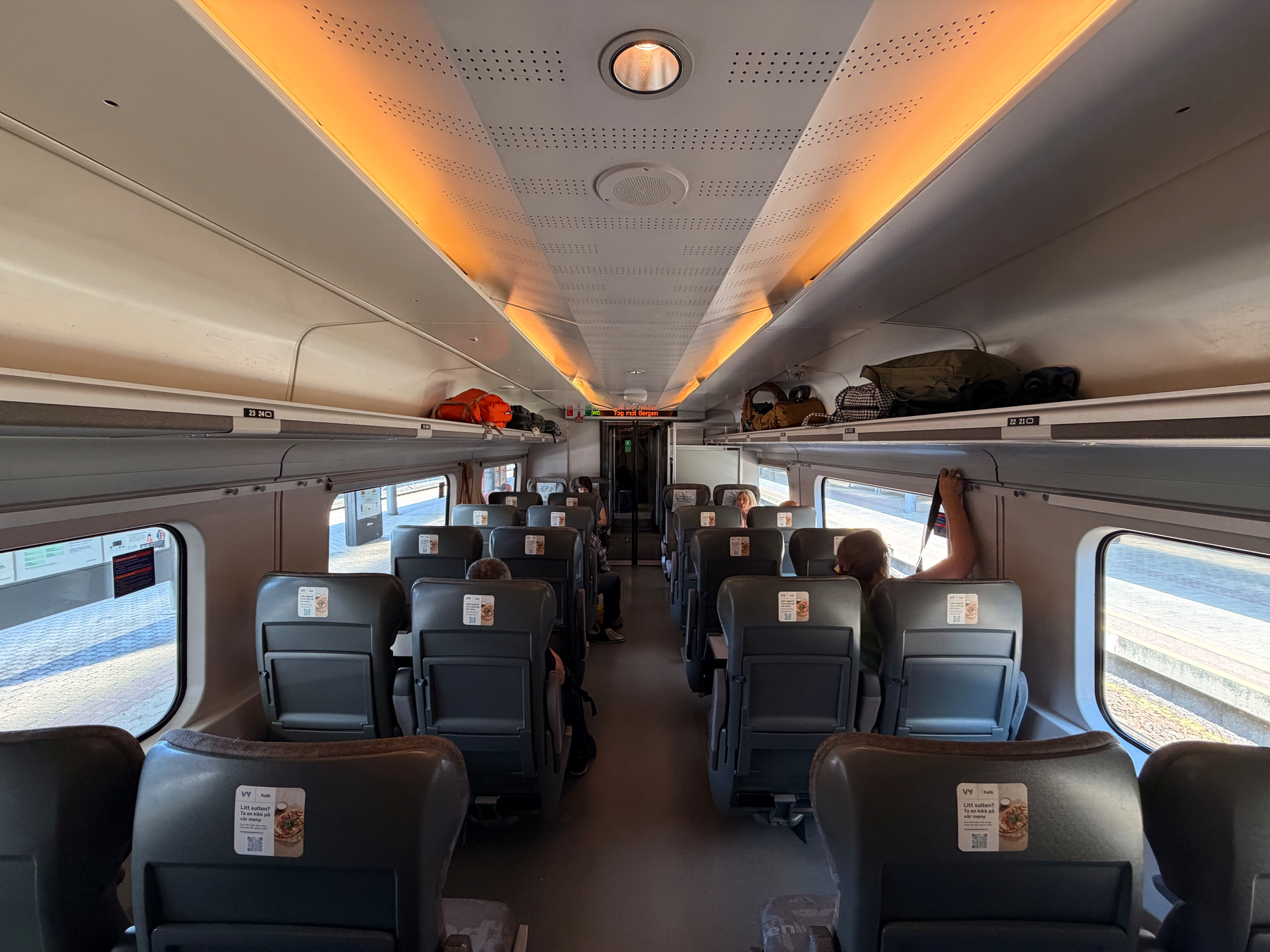 Auto-generated description: An interior view of a modern train carriage shows passengers seated with overhead luggage racks and large windows on both sides.