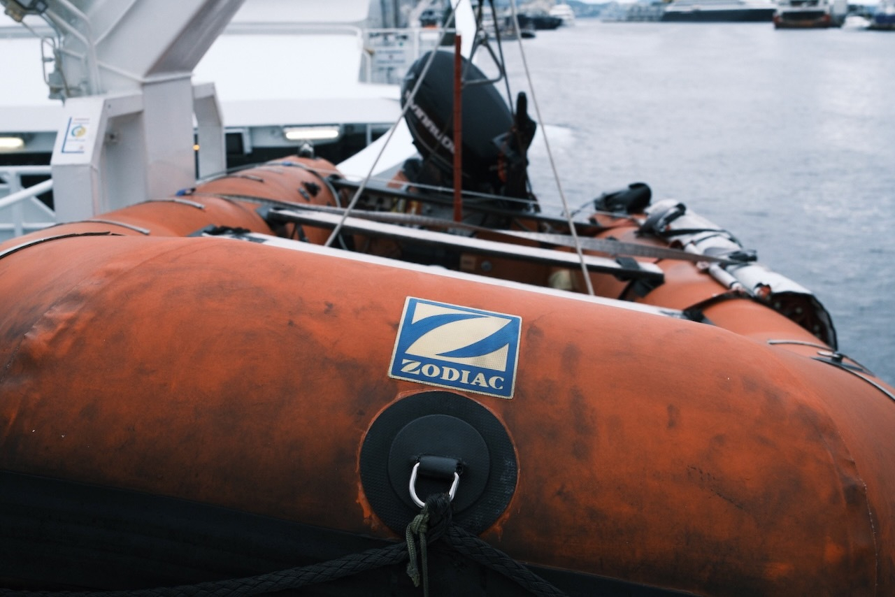 Auto-generated description: An orange Zodiac inflatable boat is secured aboard a larger vessel near a body of water, with other boats visible in the background.