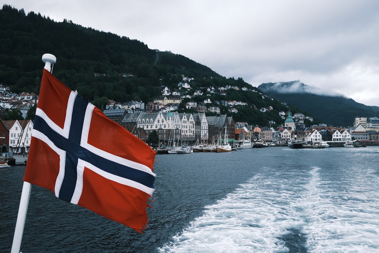 Auto-generated description: A Norwegian flag waves on a boat with a scenic view of coastal buildings and hills in the background under a cloudy sky.