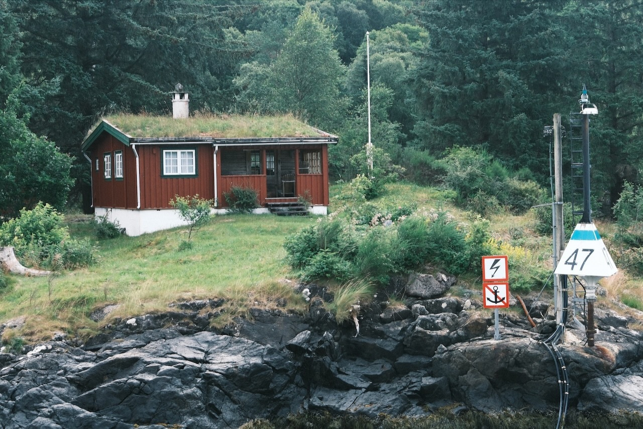 Auto-generated description: A small red cabin with a grass-covered roof is situated on a grassy area near rocky water&rsquo;s edge, surrounded by dense trees and marked with navigation signs.