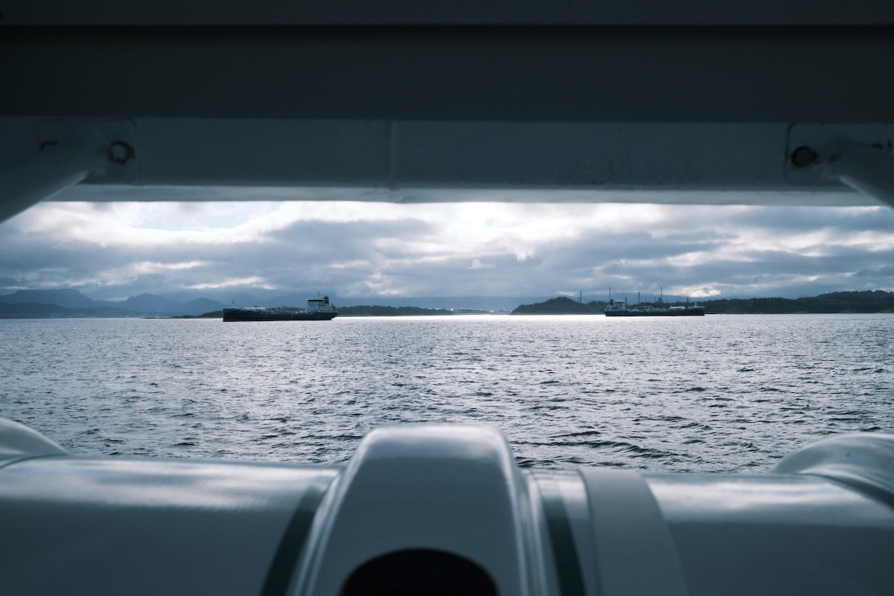 Auto-generated description: A cloudy sky is reflected over a calm sea with a distant shoreline and ships visible on the horizon, framed by the structure of a boat.