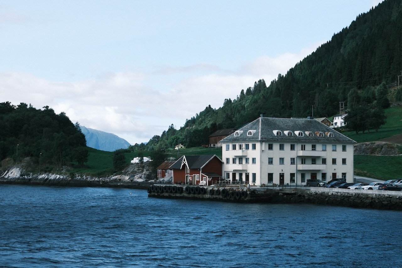 Auto-generated description: A waterfront scene features a large white building with a slate roof and several smaller red cabins nestled between lush green hills and calm blue water.